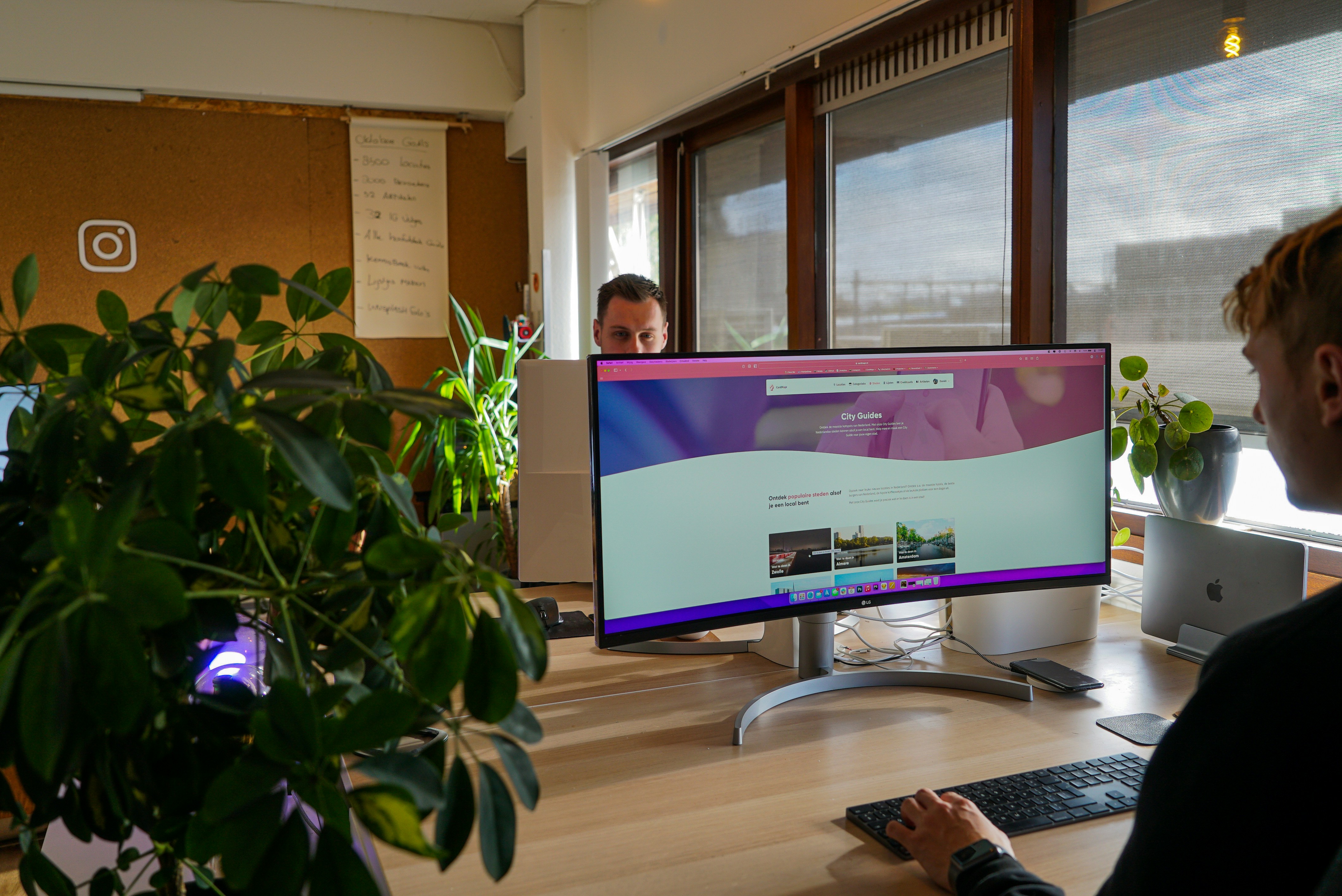 Two colleagues working in a sunlit office, one designing a website on a wide curved monitor. Photograph by CardMapr.nl.